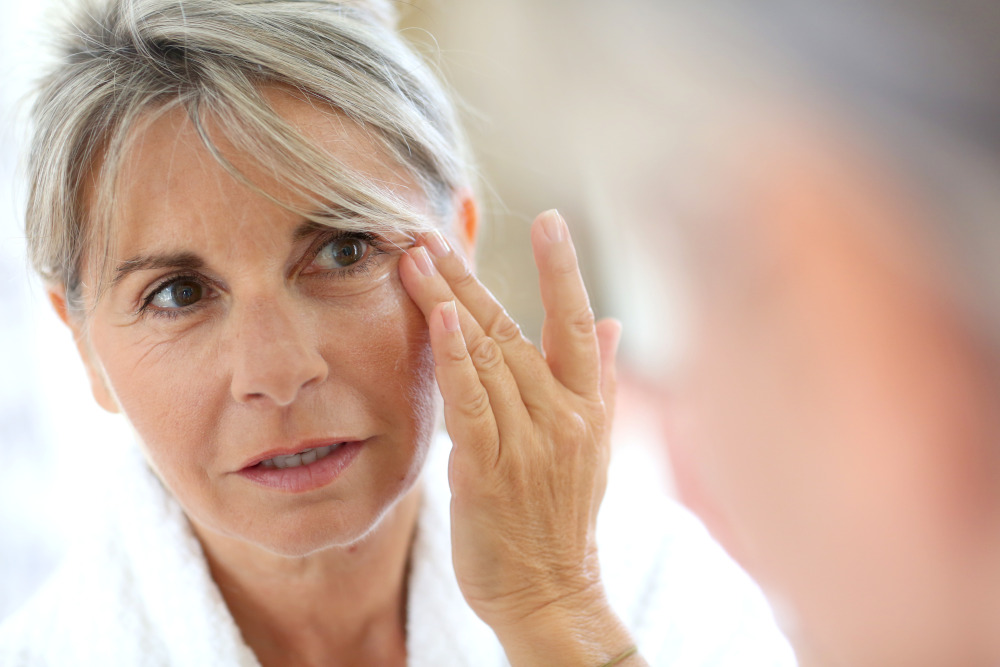 Eyelid Surgery. Woman in robe lifting her eye area with her fingers while looking at herself in the mirror.